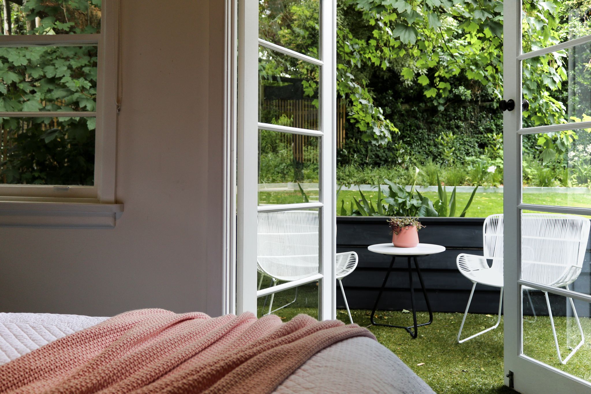 Un dormitorio con un ventanal grande, una cama con manta rosa y vistas a un jardín con sillas blancas y una mesa.