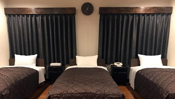 Habitación de hotel con dos camas individuales, una cama grande, cortinas oscuras, un reloj, mesitas de noche y un teléfono.