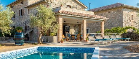 En villa i sten med en swimmingpool, utomhusmöbler och en trappa som leder upp till entrén.