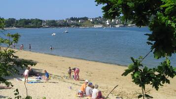Une plage où des gens se prélassent au soleil et se baignent, un rivage sablonneux et des bateaux sur l’eau.