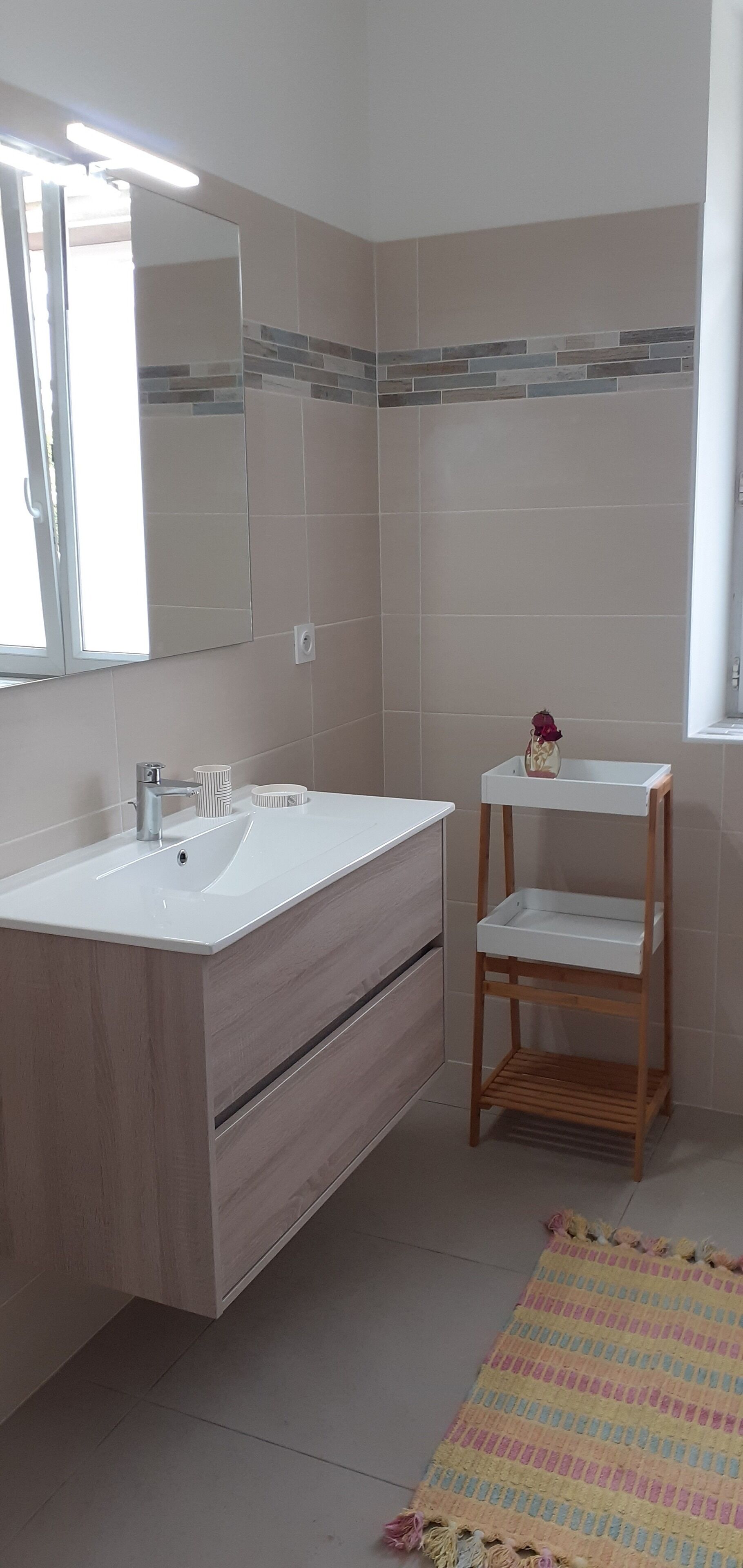 Une salle de bain moderne avec un lavabo blanc, un miroir et des murs carrelés. On y trouve une étagère en bois sur laquelle est posé un vase, ainsi qu’un tapis à rayures sur le sol.