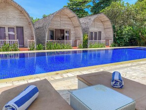 A swimming pool with a clear blue water surface, surrounded by a tiled deck. There are two small, arched-roofed structures with windows and a door, likely part of the hotel. - Villa Del Mar Canggu (Canggu)