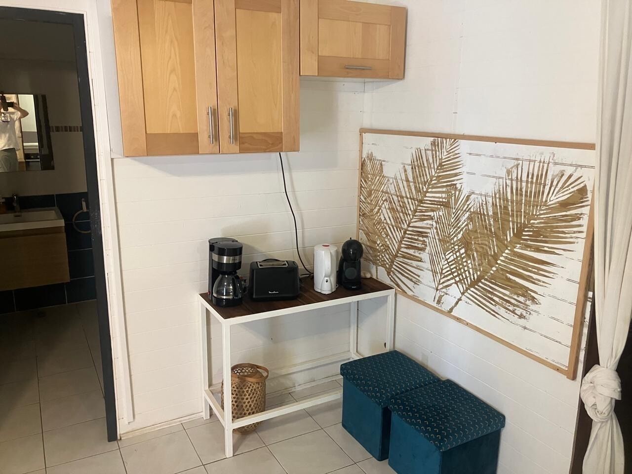 A compact kitchenette with a coffee maker, toaster, and a small shelf. There is a framed artwork on the wall and a bench in the corner.
