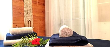 A room with a wooden wall, a macrame wall hanging, and a bed with neatly folded towels and a rolled-up towel.