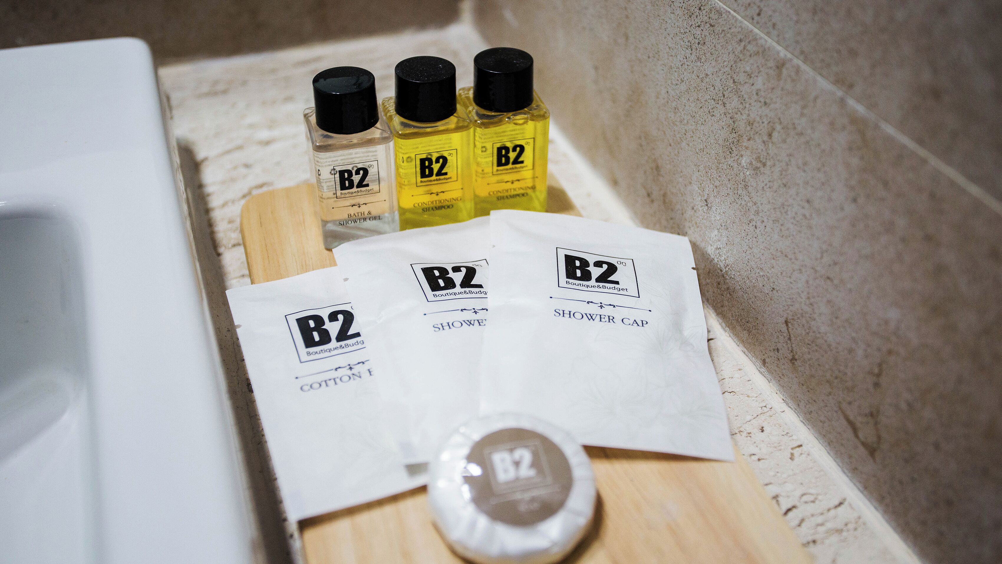 A bathroom with a bathtub, a wooden shelf holding three bottles of B2 conditioning shampoo, two shower caps, and a bottle of B2 shower gel.