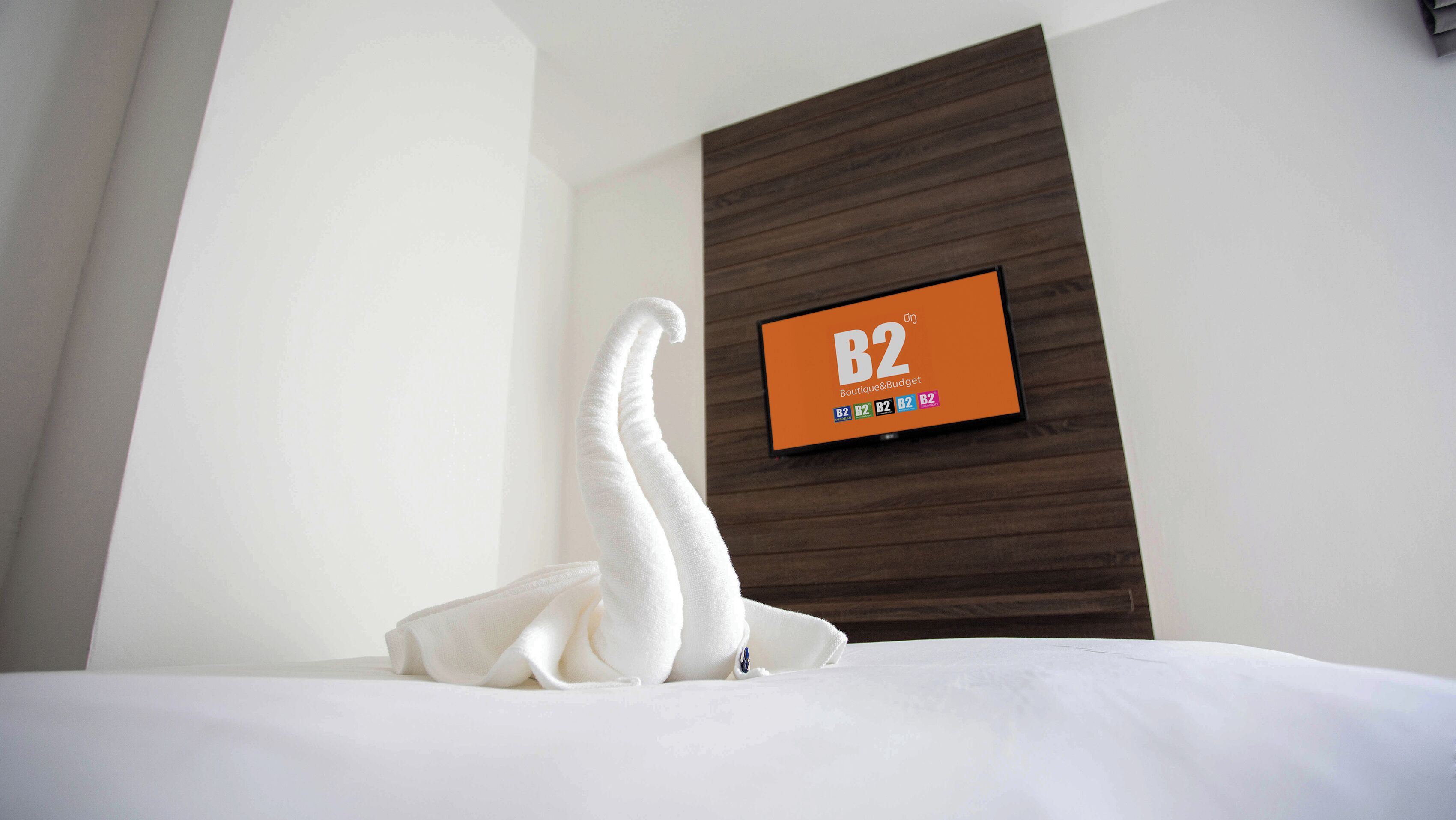 A hotel room with a bed, a white towel, and a wall-mounted TV displaying 'B2 Boutique & Budget'.