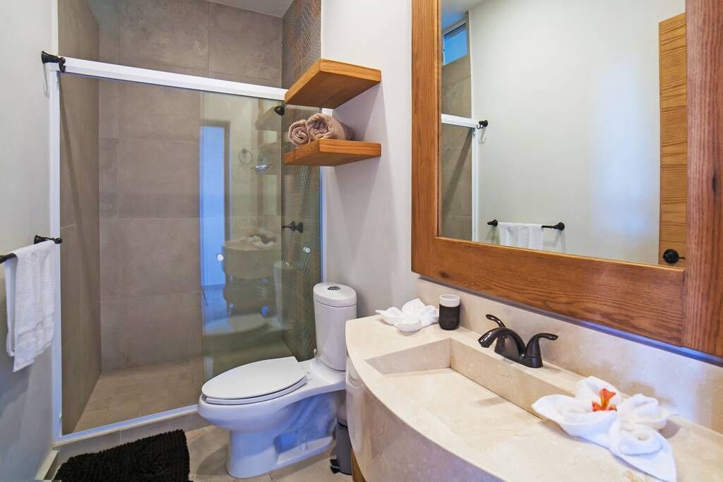 A modern bathroom with a glass-enclosed shower, a toilet, a sink with a mirror, and wooden shelves with towels.