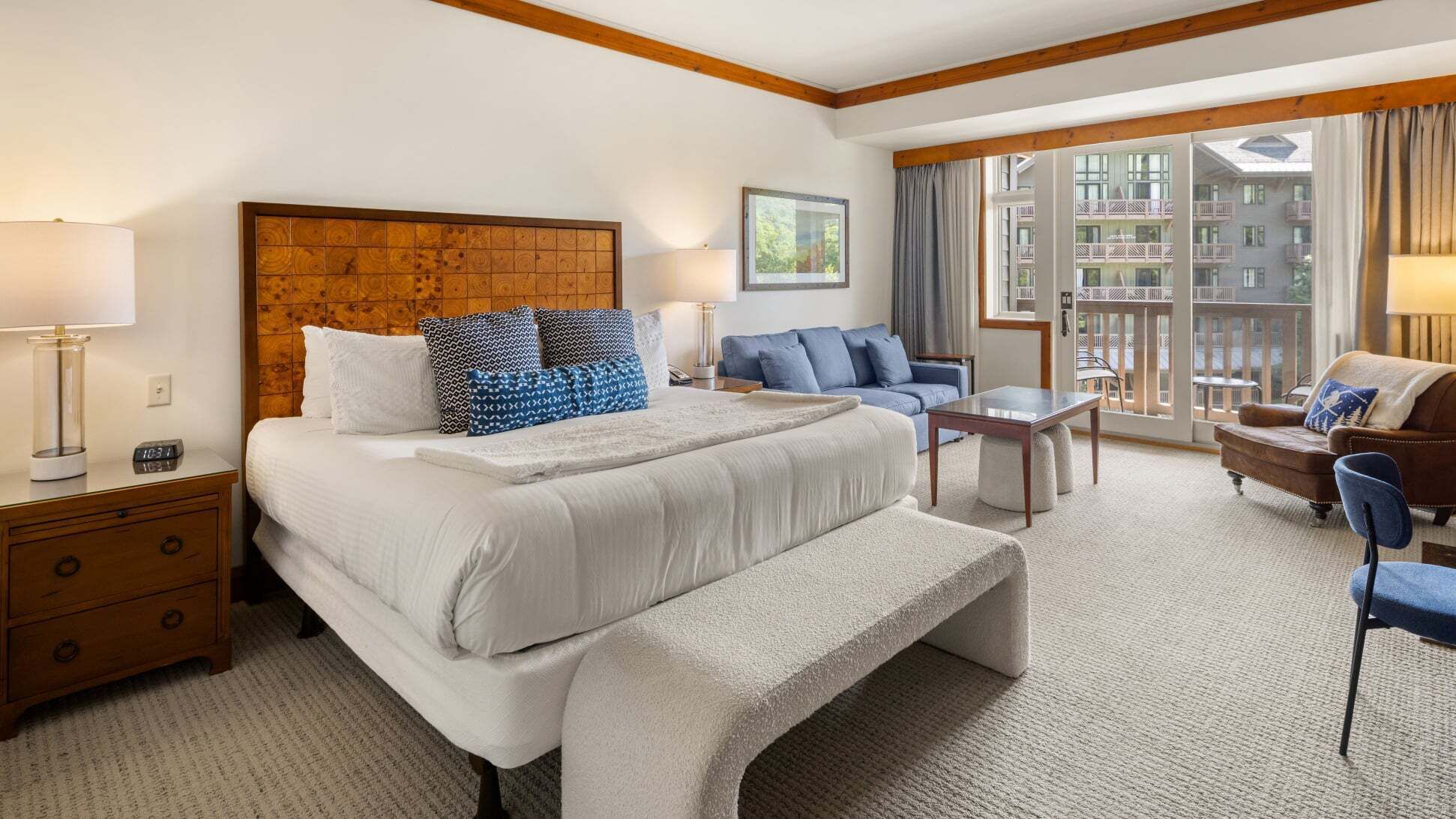A hotel room with a large bed, a desk, a chair, a sofa, and a balcony with a view of buildings.