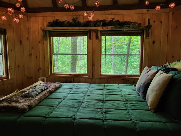 Une pièce chaleureuse avec un lit vert, deux fenêtres donnant sur une forêt, des murs en bois et des guirlandes lumineuses.