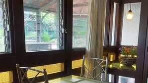 Comedor con mesa de vidrio y sillas de metal, una mesita auxiliar con una planta y vistas al exterior con vegetación.