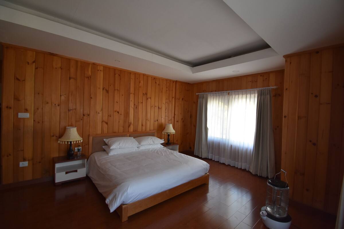 A bedroom with wooden panel walls, a large bed, two bedside tables with lamps, a window with curtains, and a water dispenser.
