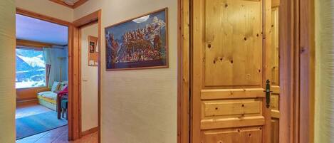 A hallway with a wooden door, a painting of a snowy landscape, and a bed with a view of the outdoors.