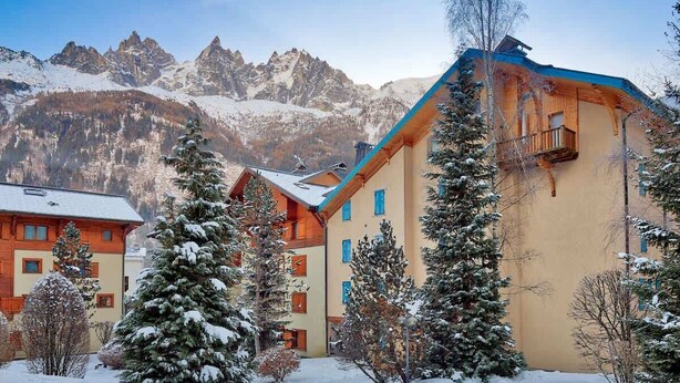 A snow-covered mountain resort with multi-story buildings, including a prominent building with a blue roof and a balcony. - Petit Jardin 2 Bed Patio Apt Central Chamonix (Chamonix-Mont-Blanc)