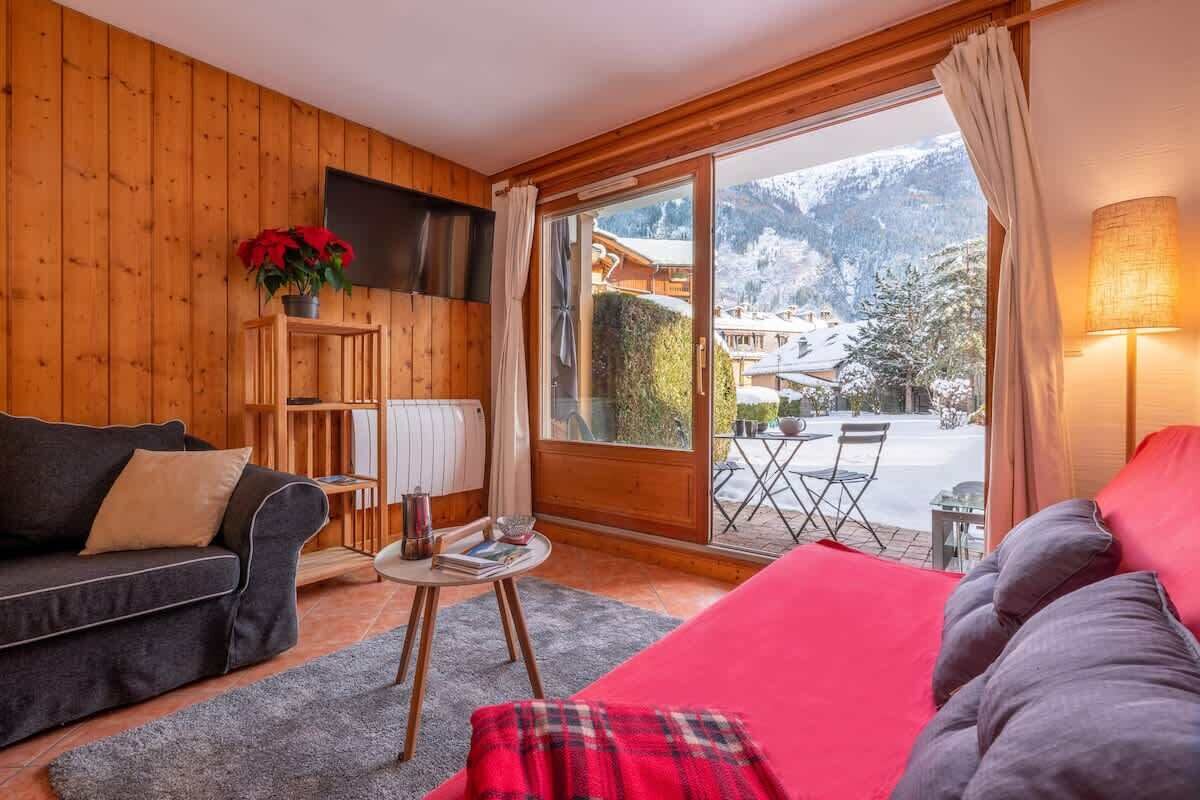 A cozy room with a wooden interior, a sofa, a red bed, a small table with books, and a view of a snowy mountain landscape through a glass door.