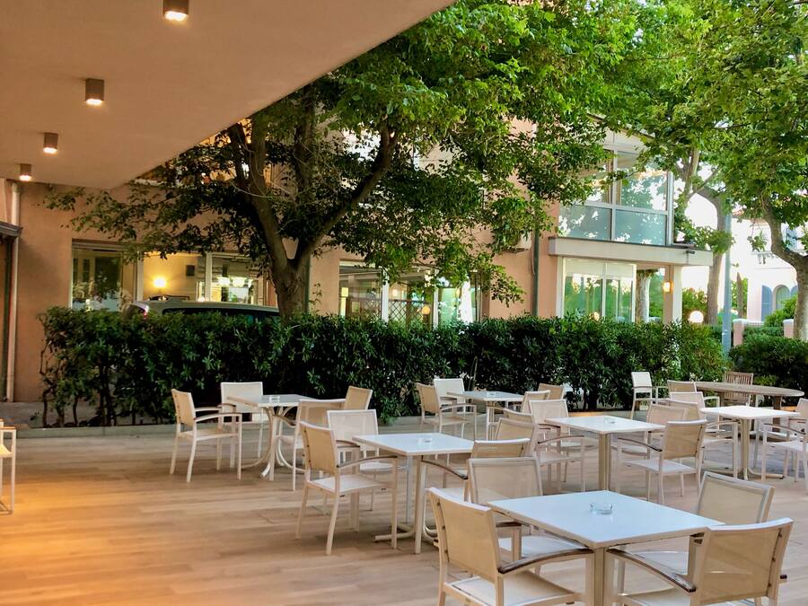 Un café al aire libre con mesas y sillas, rodeado de follaje y un edificio al fondo.