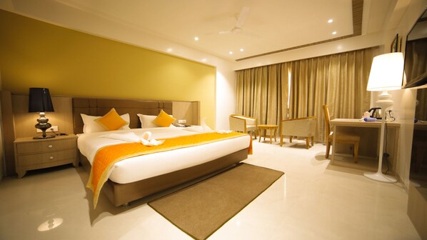 A hotel room with a large bed, a desk, a chair, a nightstand, a lamp, and a ceiling fan. - Dhamma Grand Hotel Resort (Gaya)