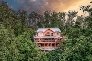 A two-story wooden house surrounded by dense forest.