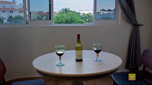 A round table with a bottle of wine and two glasses, positioned by a window with a view of buildings and trees. - "Marietas", One bedroom Apartment. You can walk to Beach and Restaurants.  (Punta de Mita)
