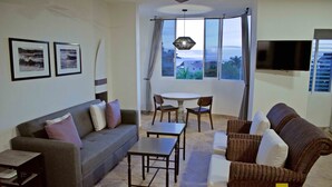 A living room with a grey sofa, a brown wicker sofa, a small table, and a dining table with chairs. There is a TV mounted on the wall and a window with curtains. - "Marietas", One bedroom Apartment. You can walk to Beach and Restaurants.  (Punta de Mita)