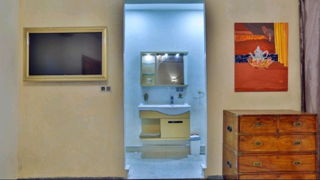 A room with a television on the left, a bathroom with a sink and mirror, and a wooden dresser on the right.