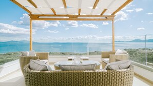 Un balcón con mobiliario de mimbre, una mesa de vidrio y vistas al mar y a las montañas.