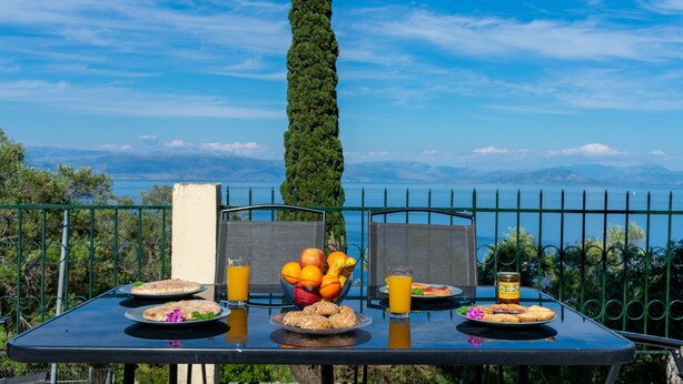 Mesa posta ao ar livre com pratos de comida, uma tigela de frutas e copos de suco, com vista para uma paisagem deslumbrante.