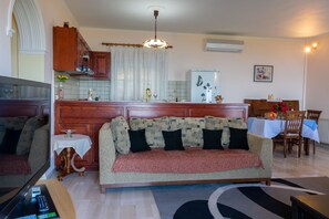 A living room with a sofa, a TV, a small table with a vase, and a kitchen area with a refrigerator and cabinets. - Villa Emily (Corfu)