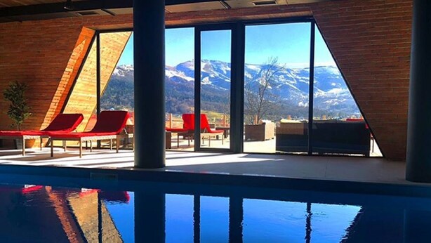 A modern indoor pool area with red lounge chairs, large windows offering mountain views, and a clear blue pool.
