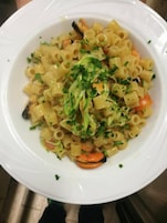 En vit tallrik med pasta, grönsaker och örter.