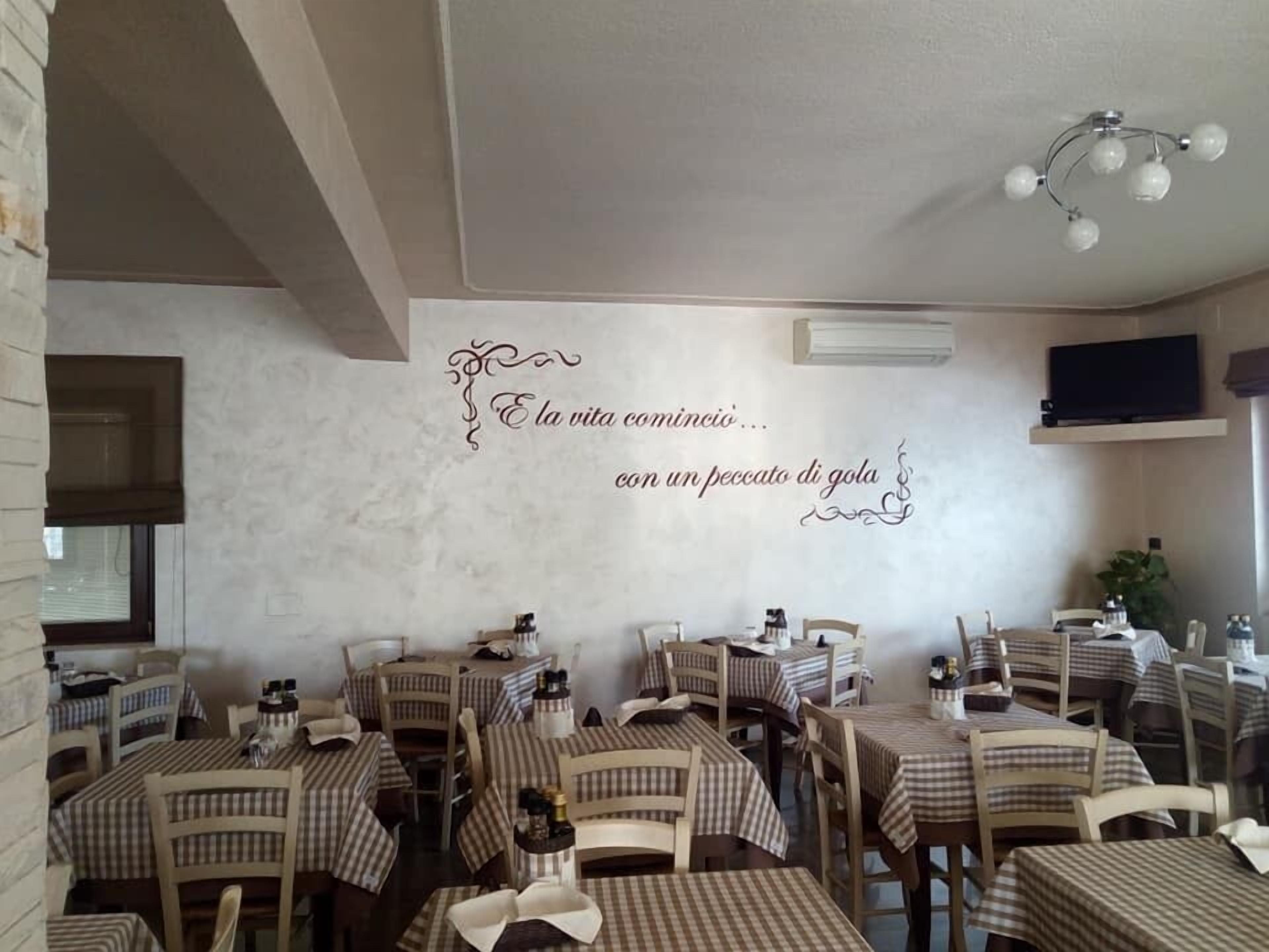 L’intérieur d’un restaurant avec des nappes à carreaux, des chaises en bois et un mur décoratif orné de texte en italien.