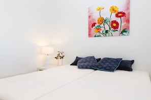 A bedroom with a bed, pillows, a nightstand, a lamp, and a painting of flowers on the wall.