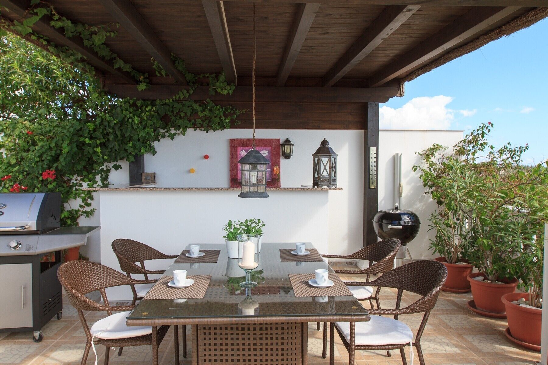 An outdoor patio with a glass table, wicker chairs, a grill, and potted plants.