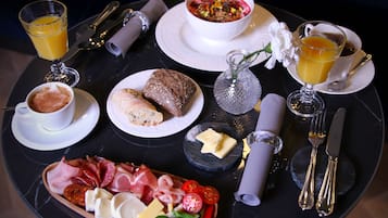 Una colazione con diversi cibi, tra cui pane, formaggi, salumi e un vassoio di frutta, disposti su un tavolo rotondo.