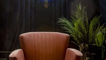 Un sillón rosa y mullido en una habitación con poca luz, con un reloj grande en la pared y una planta en maceta a un lado.