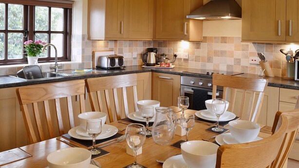 Een keuken met houten kastjes, een eettafel voor zes personen gedekt met witte borden en glazen, en een raam met uitzicht op groen.
