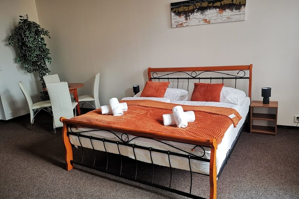 A neatly made bed with a metal frame, white bedding, and orange accents, accompanied by a small bedside table and a wall-mounted plant.