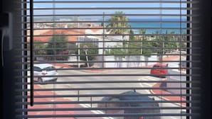 Une fenêtre avec des stores horizontaux, donnant sur une zone côtière avec des bâtiments et des voitures garées.