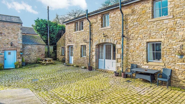 A stone building with a cobblestone courtyard, a wooden door, and a table with chairs.