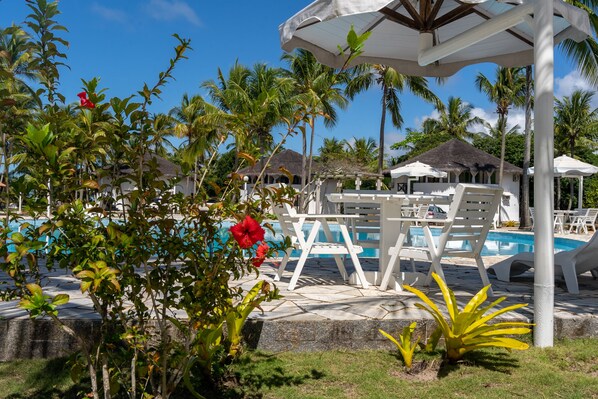 Un área de piscina con sillones blancos y una zona de estar cubierta, rodeada de palmeras y plantas tropicales.