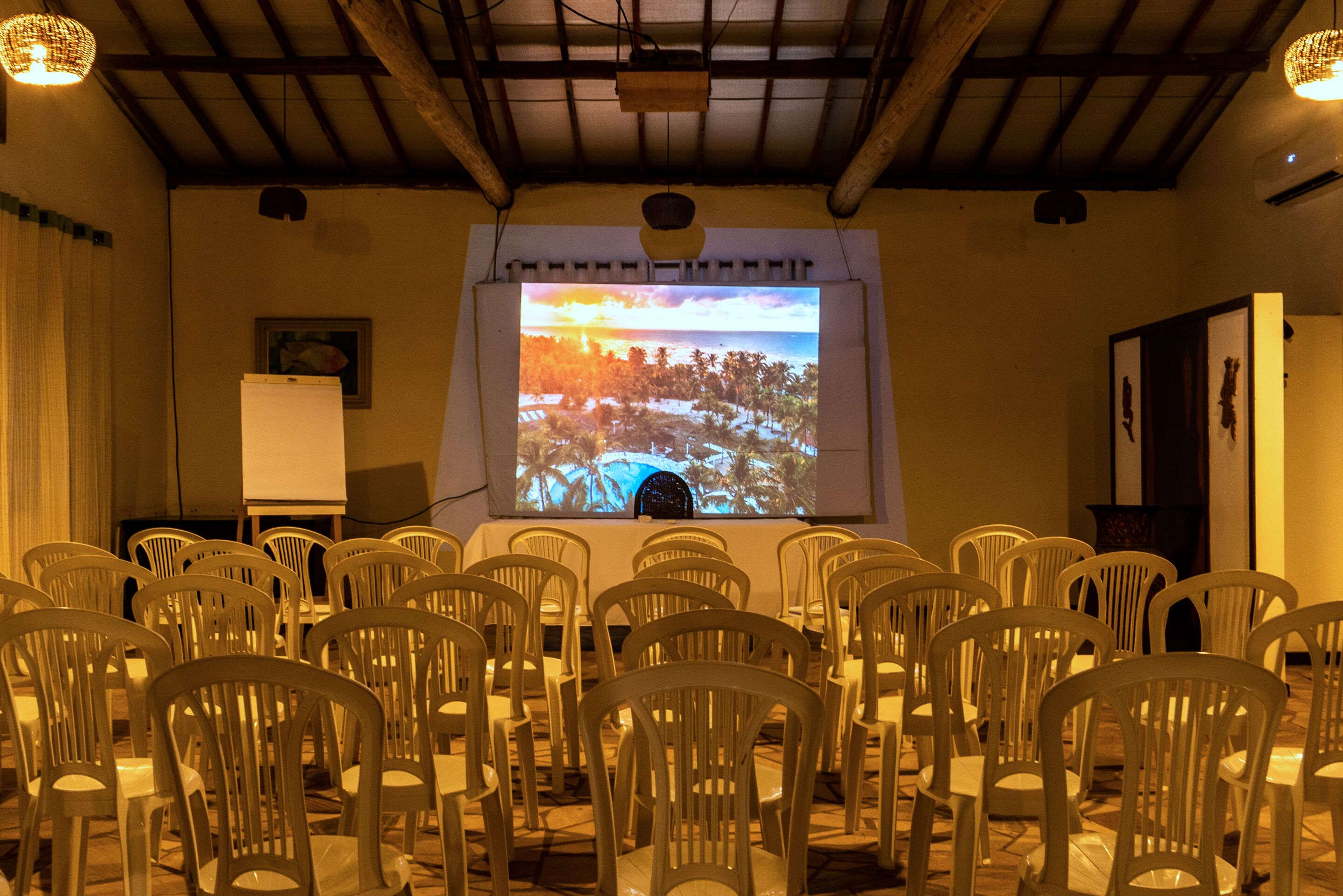 A conference room with rows of white plastic chairs, a projection screen displaying a tropical scene, and a whiteboard.