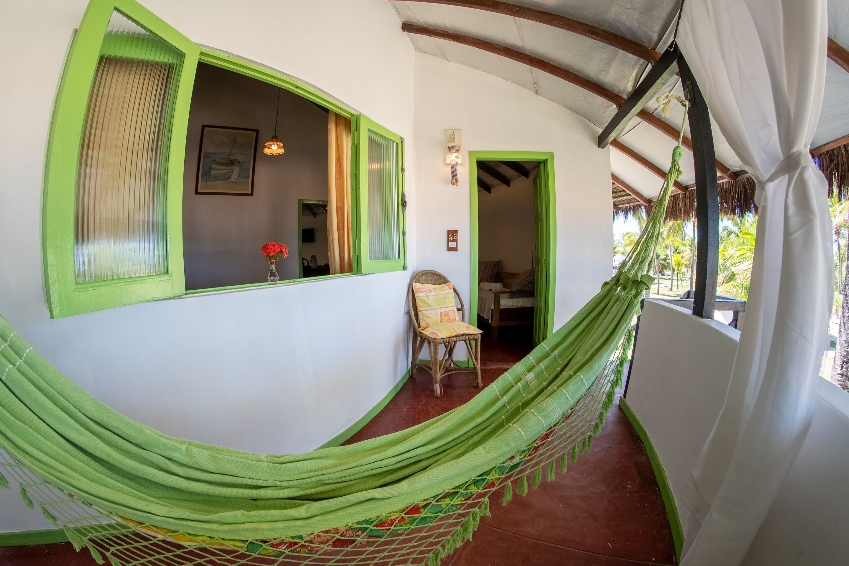 Una habitación con una hamaca verde, una silla de mimbre, una ventana con contraventanas verdes y vistas a palmeras afuera.