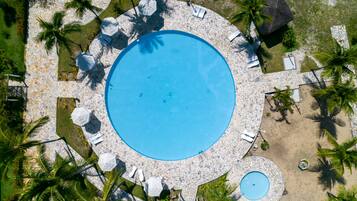 An aerial view of a circular swimming pool surrounded by lounge chairs and umbrellas, with palm trees and a small adjacent pool.