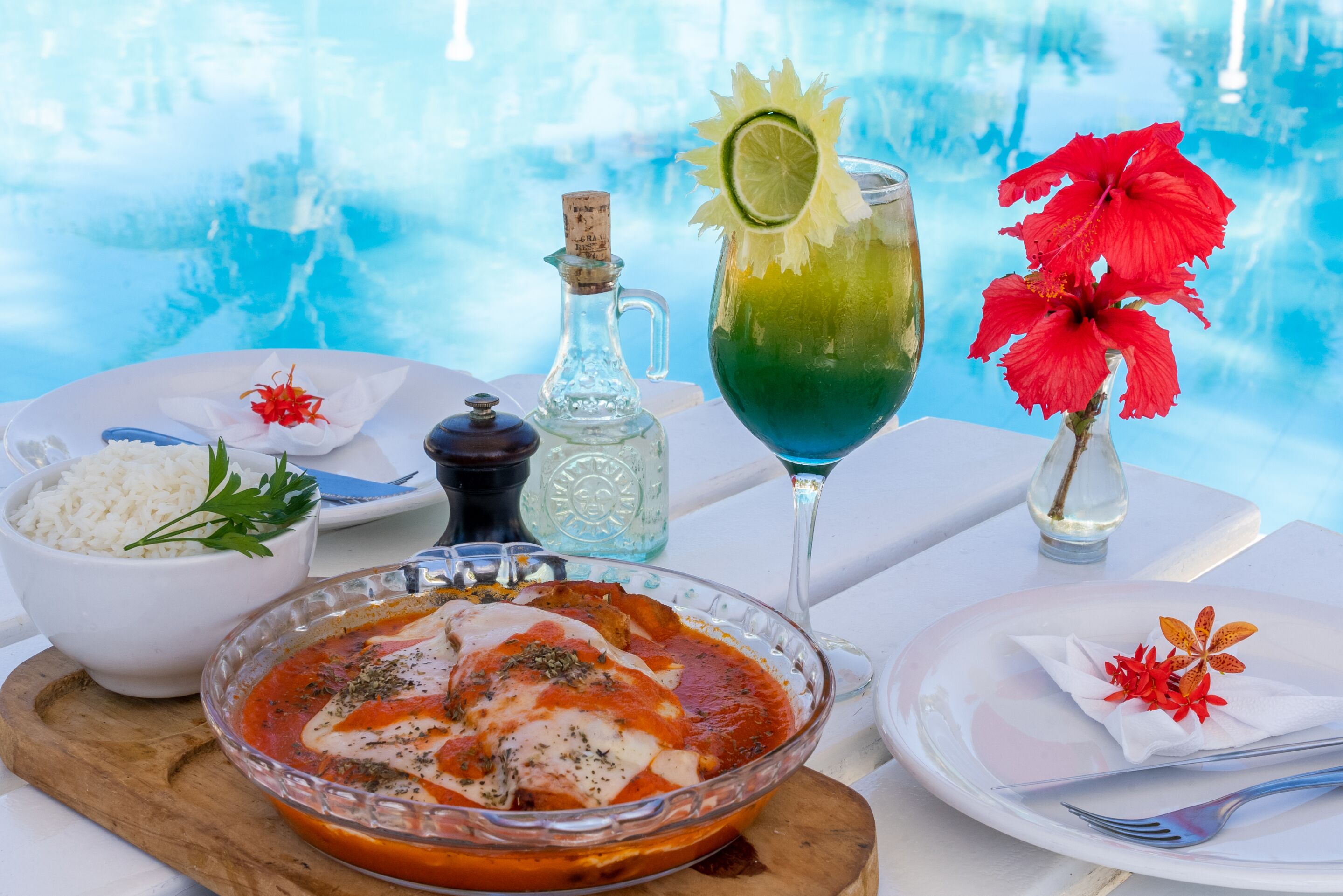 A dining table with a bowl of rice, a plate of food, a glass of drink, and a vase with flowers.