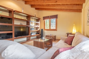 A cozy living room with a sofa, a glass coffee table, wooden shelves, a TV, and a window with a view. - Fantastic Finca with Wi-Fi, Air Conditioning, Garden and Terrace (Algaida)