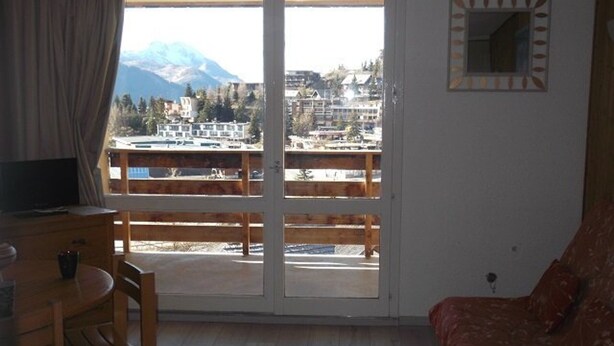 A room with a wooden desk, a chair, a sofa, and a large window with a view of buildings and mountains. - Cozy 4-Person Studio, Ski-In/Ski-Out, South-Facing Balcony, Modern Amenities (Orcières Merlette)
