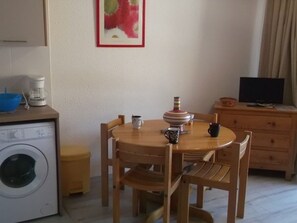 Um espaço de estar compacto com uma mesa de jantar redonda de madeira, uma máquina de lavar, uma cafeteira e uma televisão sobre um móvel de madeira.
