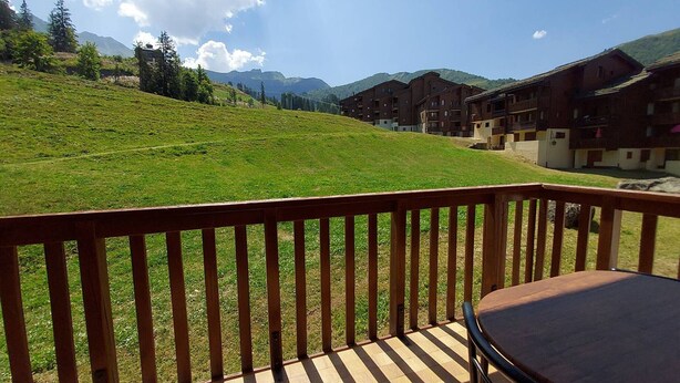 Une terrasse en bois avec une table, offrant une vue sur une colline verdoyante et une rangée de bâtiments.