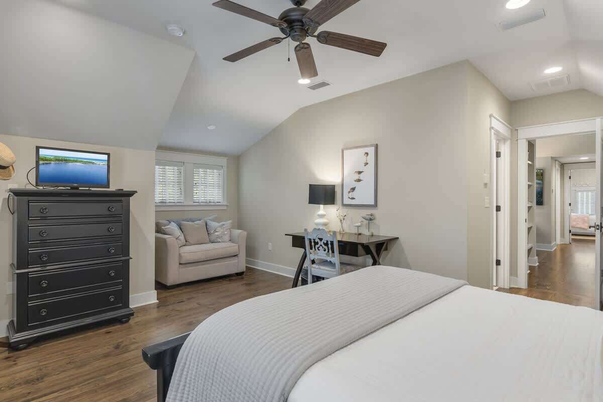 A bedroom with a bed, a dresser, a desk, a chair, a ceiling fan, a TV, and a window.