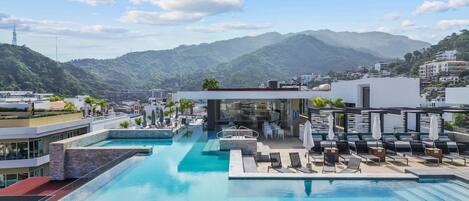 A rooftop infinity pool with lounge chairs and a bar area, overlooking a mountainous landscape.