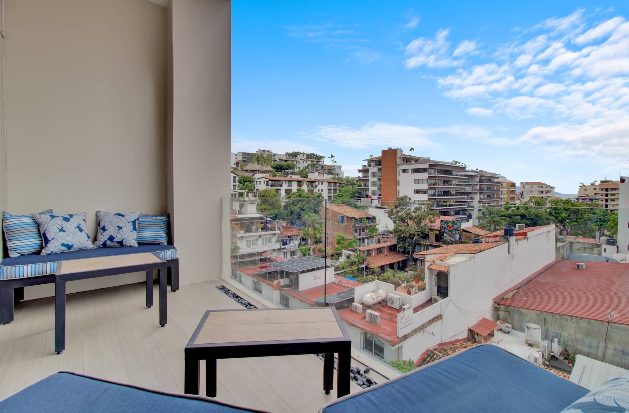 En balkon med en sofa, et lille bord og udsigt over bybilledet med bygninger og grønne områder.